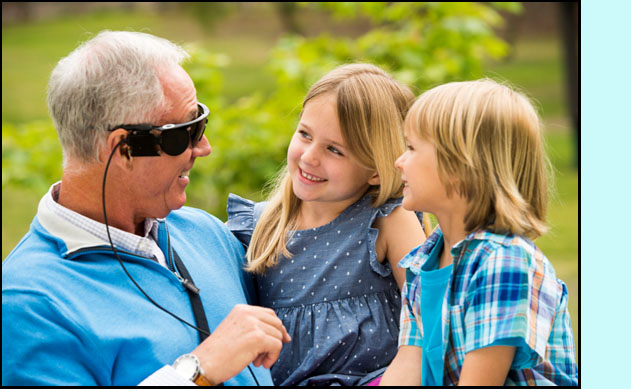 Photo shows a man smiling and looking at two children that he is holding on his lap.  He is wearing what looks like wrap-around sunglasses with a cord coming down to his shirt. 